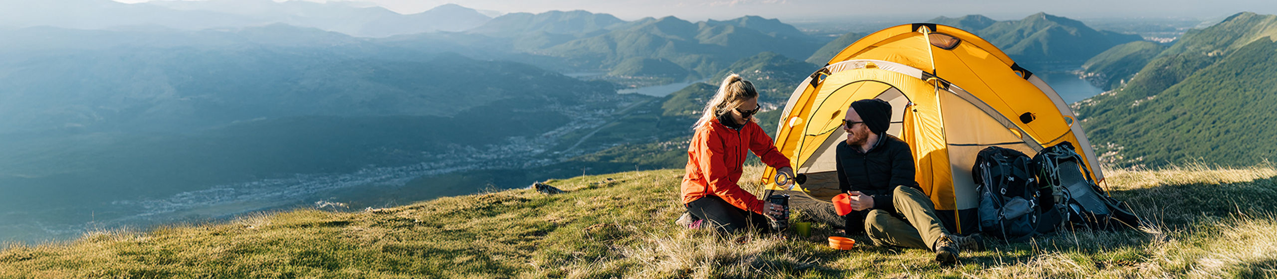 Deux campeurs sont assis à côté d’une tente jaune vif sur une crête de montagne herbeuse.