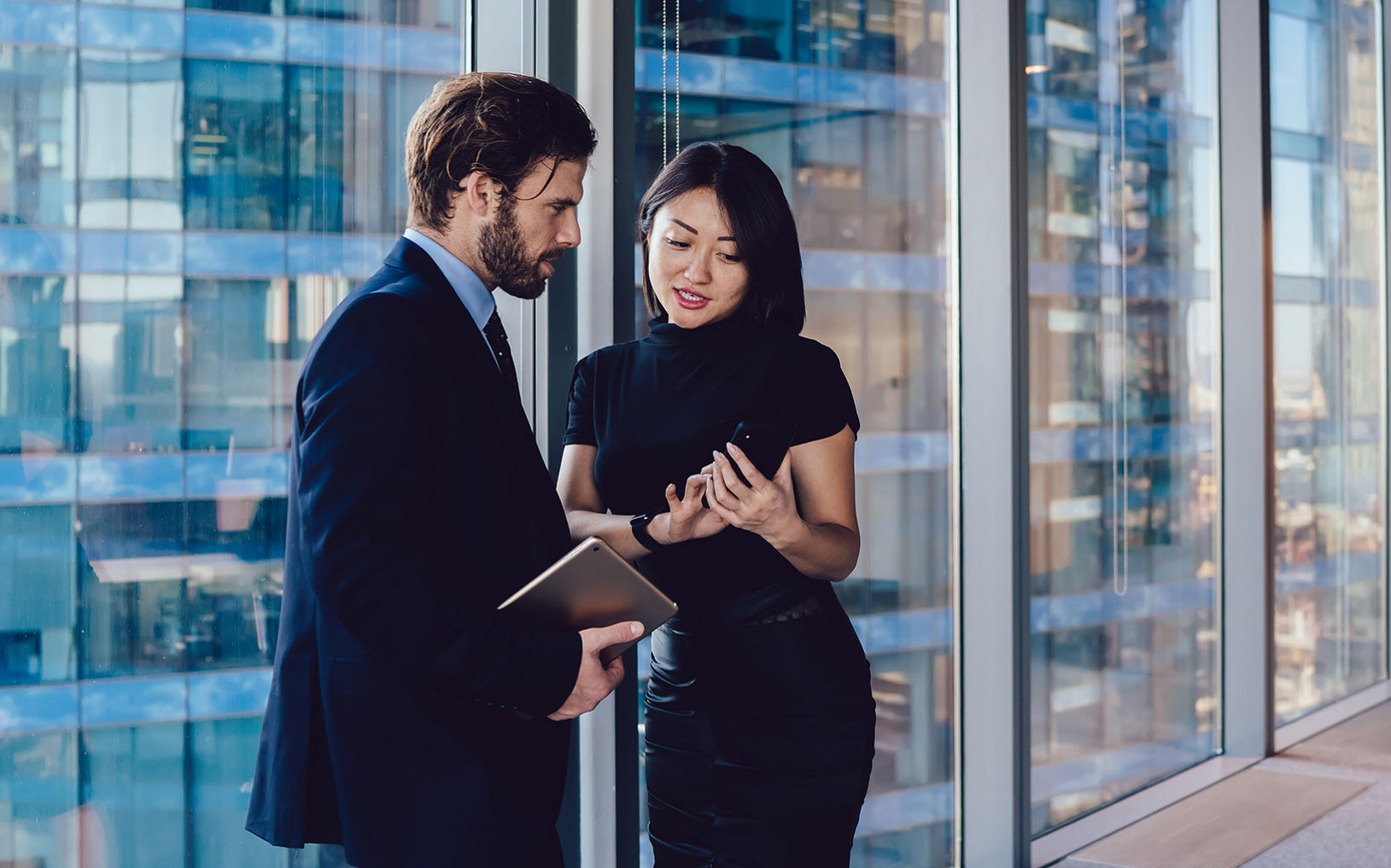 Two business professionals stand near large floor-to-ceiling windows in a modern office, engaged in a conversation. The man in a suit holds a tablet or folder while the woman in a black dress gestures with a smartphone. In the background, another colleague sits at a desk near the windows, working on a computer. The scene overlooks tall glass buildings, emphasizing a contemporary corporate environment.