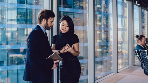 Deux professionnels en pleine conversation se tiennent près de grandes baies vitrées dans un bureau moderne. L’homme en costume tient une tablette ou un dossier, tandis que la femme en robe noire fait des gestes avec un téléphone intelligent. À l’arrière-plan, un autre collègue assis à un bureau près des fenêtres travaille sur son ordinateur. La scène donne sur de grands immeubles de verre, soulignant le caractère contemporain de cet environnement professionnel.