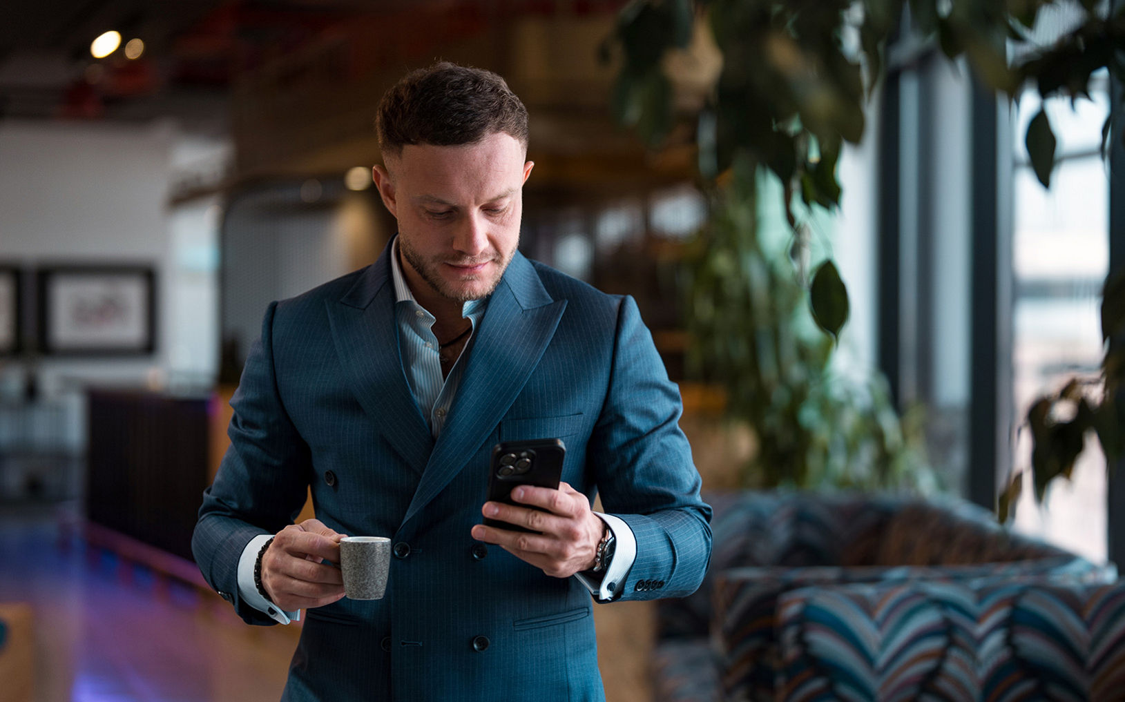 Un homme d’affaires élégamment vêtu d’un costume bleu sarcelle se tient à l’intérieur, une petite tasse de café et un téléphone intelligent à la main. Il semble consulter son téléphone tout en traversant un salon ou un bureau moderne. Les grandes baies vitrées en arrière-plan laissent entrer la lumière naturelle, tandis que les sièges à motifs et les plantes créent une ambiance élégante et contemporaine. L’ensemble de la scène évoque un mode de vie professionnel et un moment de la journée de travail bien rempli.