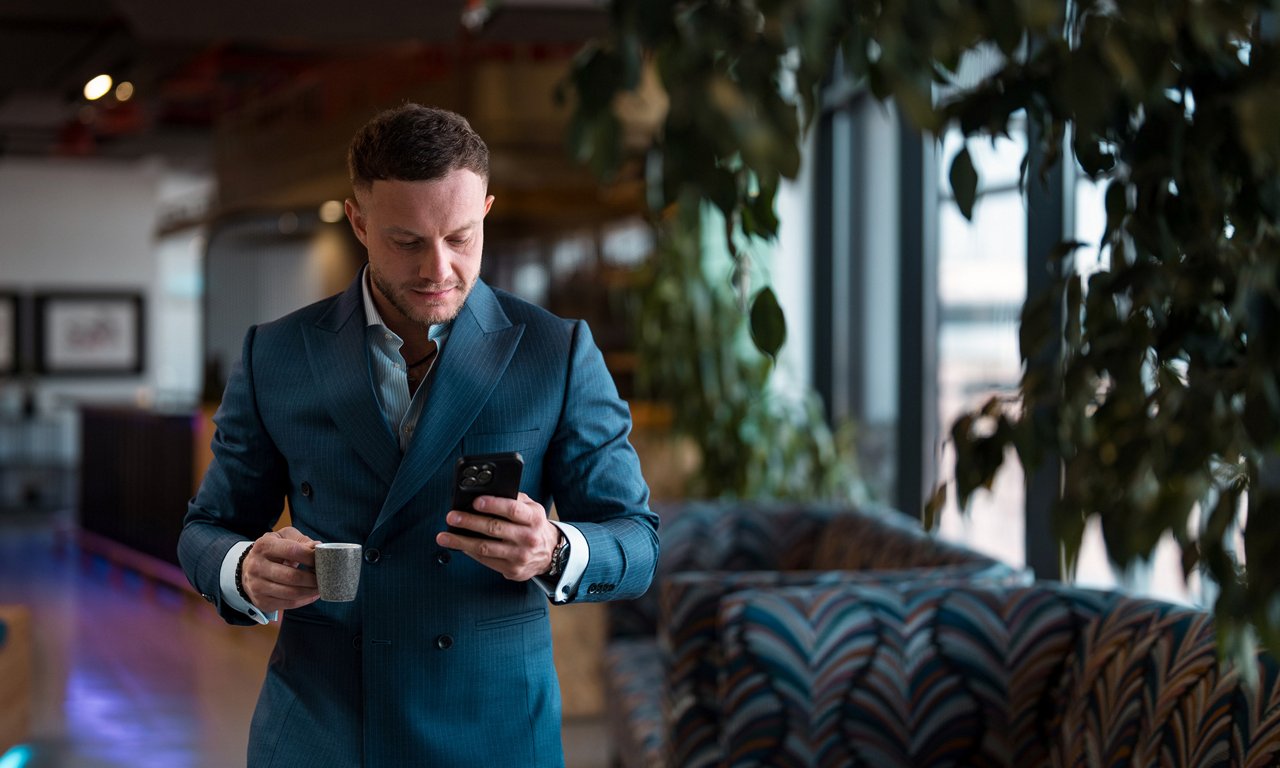 A well-dressed businessman in a teal suit stands indoors holding a small coffee cup and a smartphone. He appears to be checking his phone while walking through a modern lounge or office space. Large windows in the background let in natural light, and patterned seating and plants create a stylish, contemporary environment. The overall scene suggests a professional lifestyle and a busy workday moment.