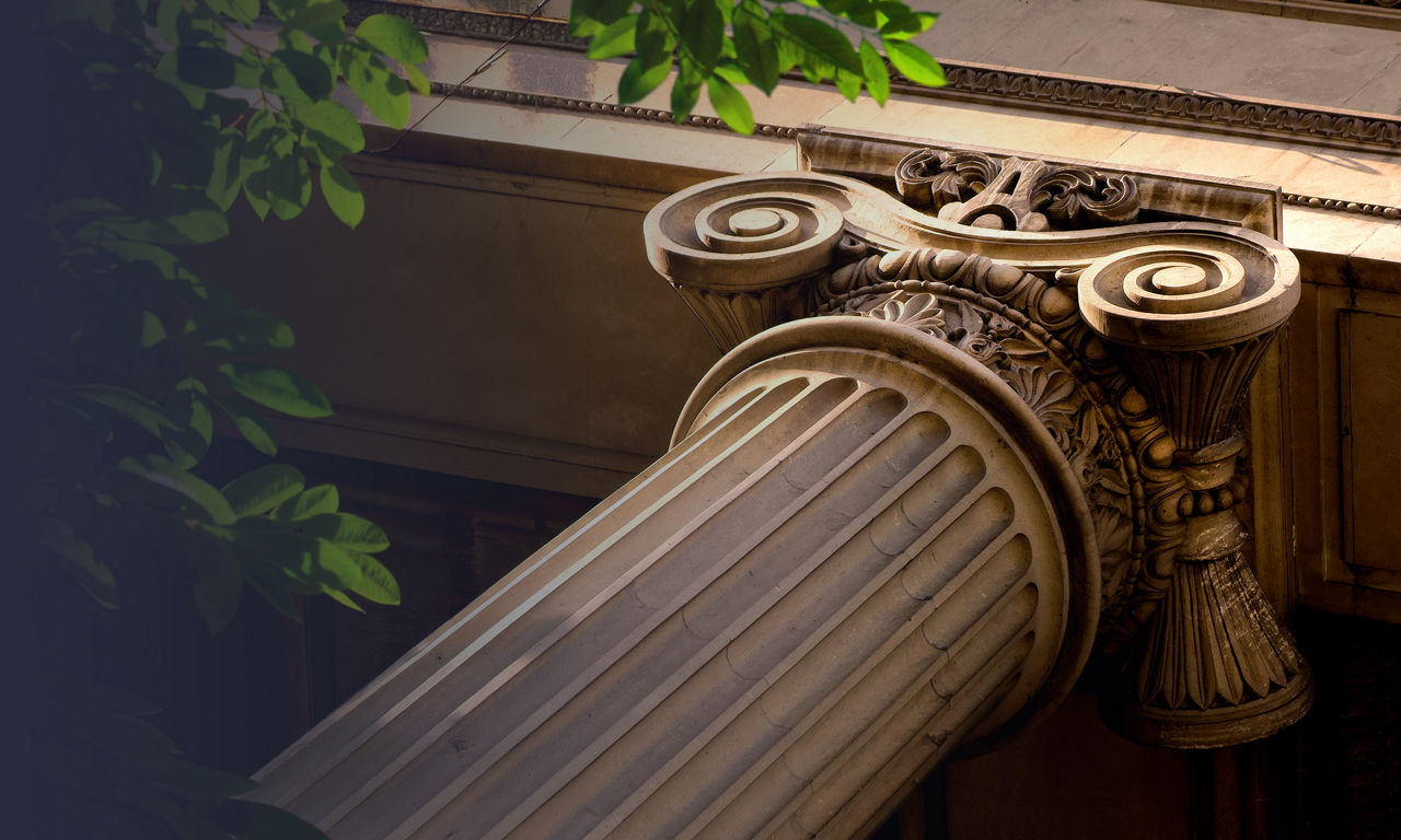 A detailed view of a classical Ionic column capital is shown in warm light. The fluted shaft leads up to ornate volutes and carved decorative elements. Architectural details of the building facade are visible in the background. Green leaves frame the scene from above, adding contrast to the stone textures.