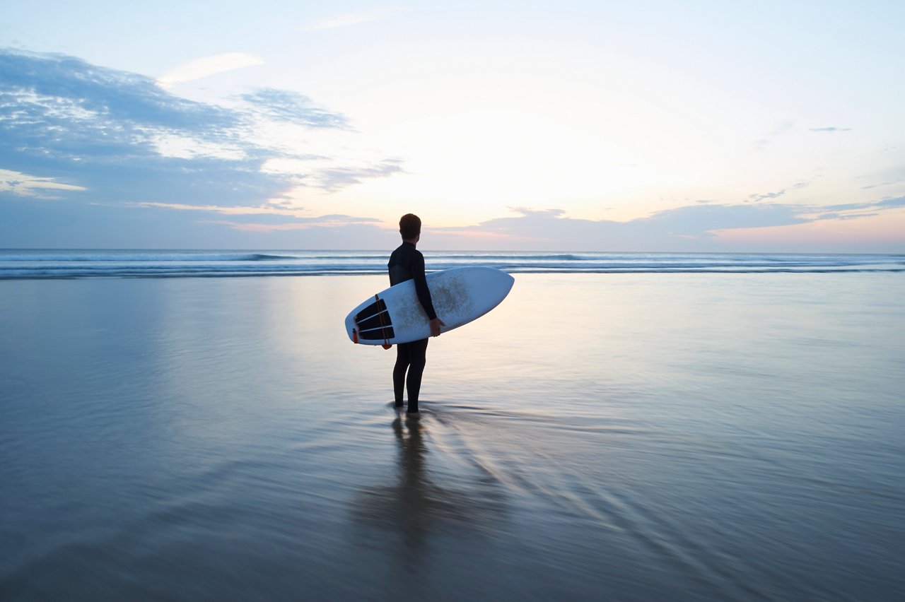 A surfer stands in the shallow waters and looks out to sea at dusk.