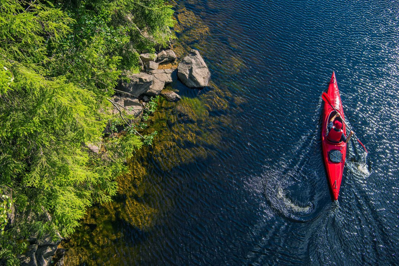 River Kayaker Aerial View. Caucasian Sportsman in the Red Kayak Paddling on the Scenic River Along the Shore.