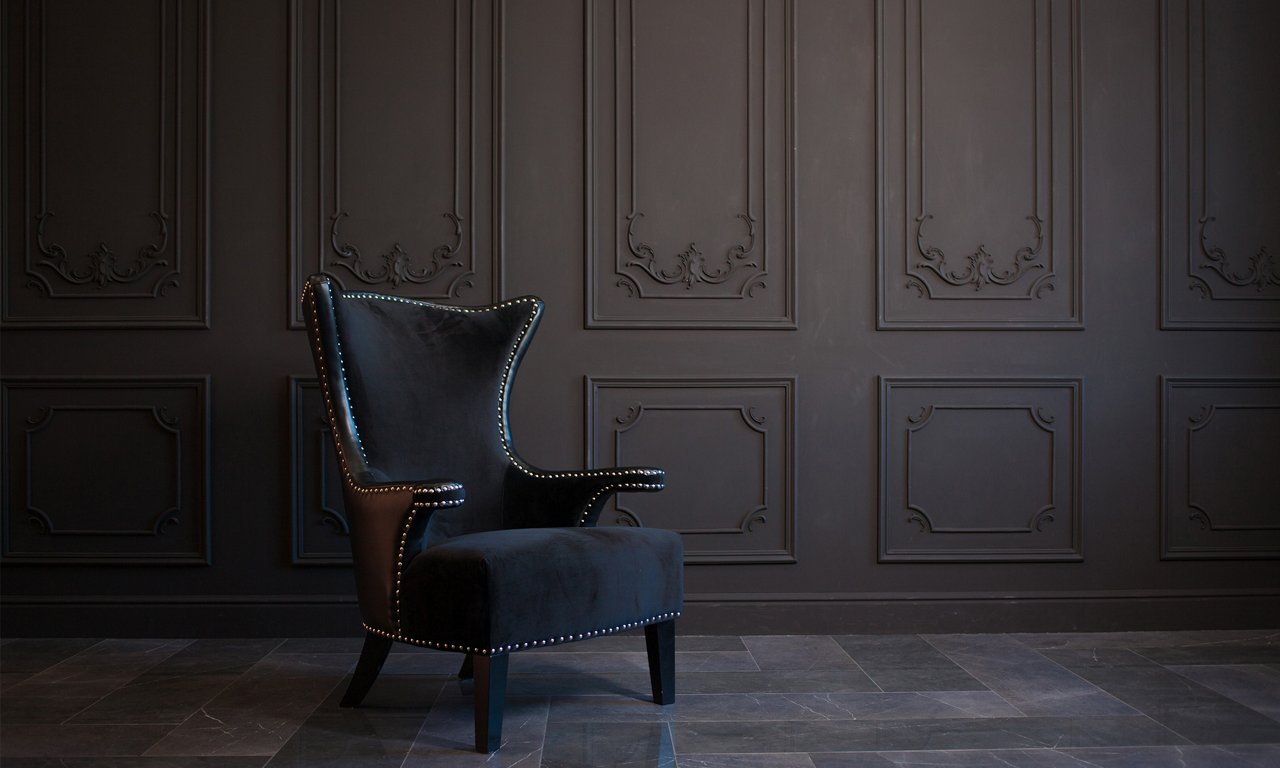 A single black upholstered armchair with studded detailing sits against an ornate dark paneled wall. The room features classic molding and a moody, minimalist interior design. Cool-toned floor tiles complement the deep charcoal and black color palette. The overall scene feels sophisticated, quiet, and luxurious.