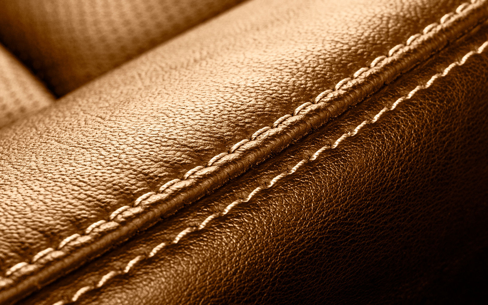 Macro photograph of brown leather upholstery with prominent stitching detail. The image focuses on a diagonal seam where two padded leather sections meet. Fine texture and grain of the leather surface are clearly visible. Warm lighting creates a rich, golden-brown tone and emphasizes the craftsmanship of the stitching.