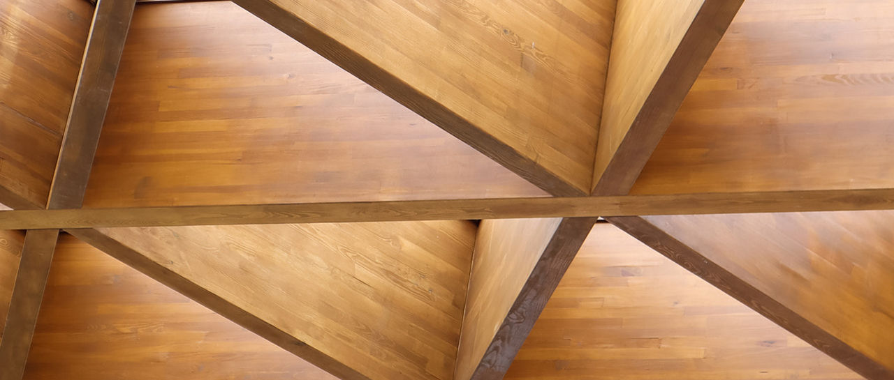 The image shows a modern wooden ceiling constructed in a repeating geometric pattern. Triangular and diamond-shaped sections form a grid across the surface. Several skylight openings allow natural light to enter through the structure. The warm wood tones and clean lines create a contemporary architectural look.