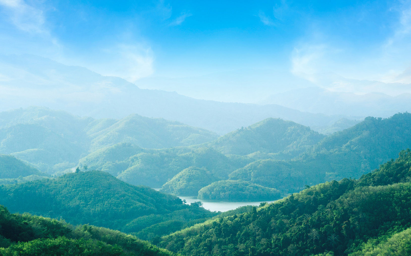 Lush green mountains stretch into the distance under a bright blue sky. Soft mist and haze create layered silhouettes of rolling hills and valleys. A narrow body of water winds between the hills, reflecting the light from above. Wispy clouds frame the upper part of the scene, enhancing the tranquil, natural atmosphere.