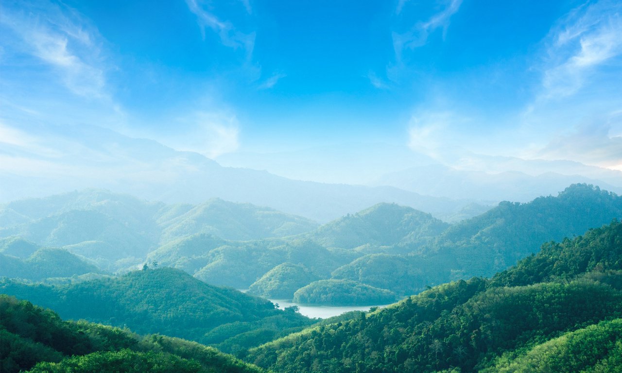 Lush green mountains stretch into the distance under a bright blue sky. Soft mist and haze create layered silhouettes of rolling hills and valleys. A narrow body of water winds between the hills, reflecting the light from above. Wispy clouds frame the upper part of the scene, enhancing the tranquil, natural atmosphere.