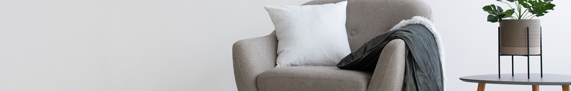 A modern gray armchair with wooden legs sits against a plain white wall in a minimalist living room. A white cushion and a draped dark gray throw blanket add softness and texture to the chair. To the right, a small round side table holds a potted green plant in a metal stand. The neutral color palette and clean lines create a calm, contemporary interior atmosphere.