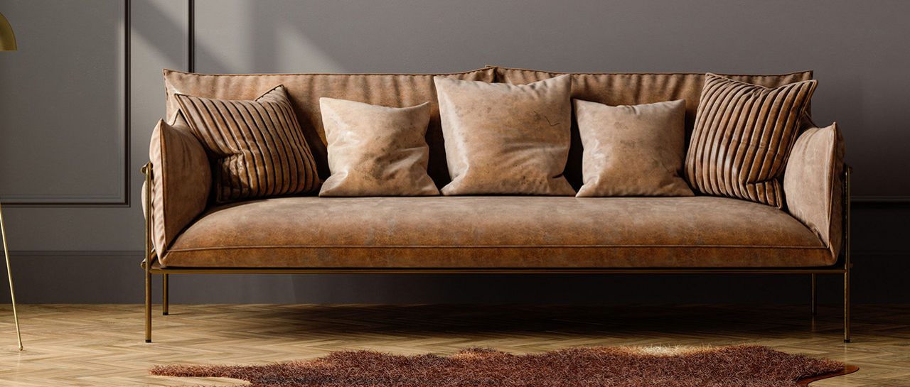 A contemporary living room features a sleek brown sofa with multiple matching cushions arranged neatly. The sofa sits against a large, paneled wall painted in a muted taupe or gray tone. A gold floor lamp with a dome-shaped shade stands to the left, while a small side table with glassware is on the right. Warm wooden flooring and a soft rug add texture and comfort to the minimalist interior design.