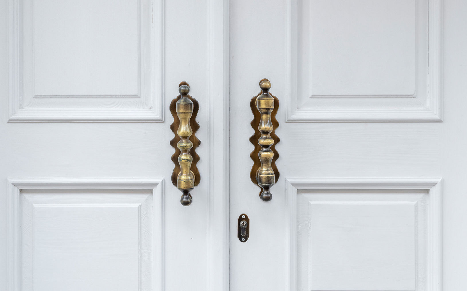 A pair of white double doors features two large, ornate brass handles. The handles have a sculpted, wavy design that stands out against the smooth, paneled door surface. A small keyhole is visible between the doors, emphasizing the entryway function. The overall scene feels clean and elegant with a minimalist architectural style.