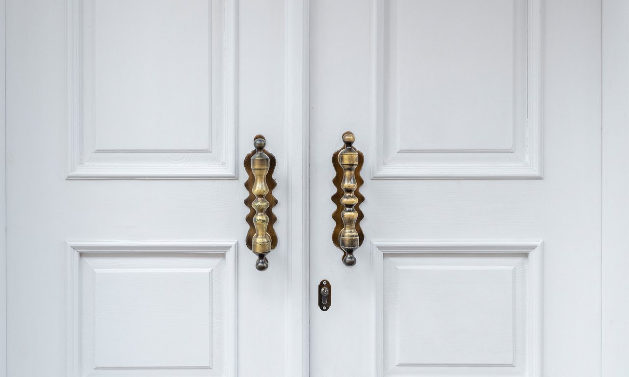 A pair of white double doors features two large, ornate brass handles. The handles have a sculpted, wavy design that stands out against the smooth, paneled door surface. A small keyhole is visible between the doors, emphasizing the entryway function. The overall scene feels clean and elegant with a minimalist architectural style.