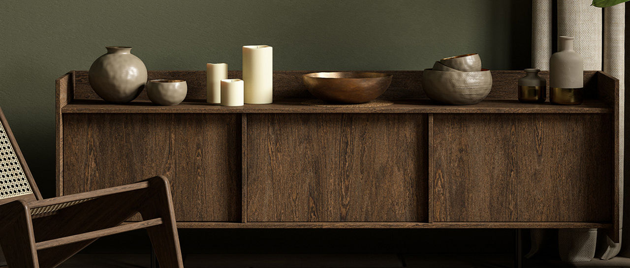A minimalist living room features a low wooden sideboard against an olive green wall. A woven lounge chair sits in the foreground beside a round patterned rug. Ceramic vases, bowls, and pillar candles are arranged on the cabinet for a natural, sculptural look. Floor-length curtains and a tall potted plant add softness and greenery to the calm, neutral space.