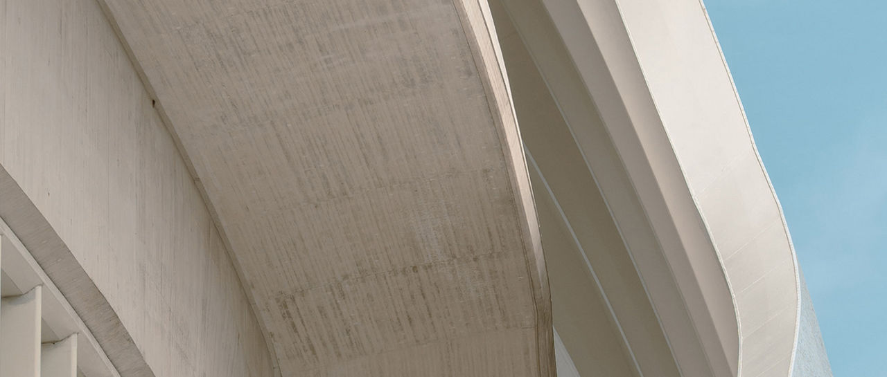 Gros plan sur la façade d’un immeuble moderne en béton aux courbes gracieuses. Les lamelles verticales dessinent un motif rythmé le long du bord de la structure. Les surfaces pâles, d’un blanc cassé, offrent un contraste tout en douceur avec le ciel d’un bleu limpide. Cette composition accentue le minimalisme, la géométrie et le design architectural contemporain.