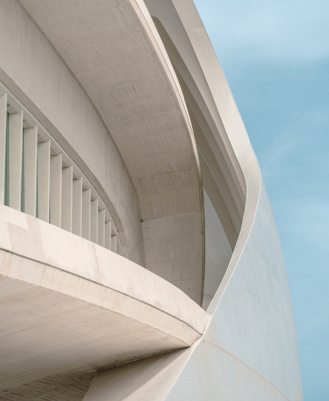 Close-up view of a modern concrete building facade with sweeping curved lines. Vertical slats create a rhythmic pattern along the structure’s edge. The pale, off-white surfaces contrast softly with a clear blue sky. The composition emphasizes minimalism, geometry, and contemporary architectural design.