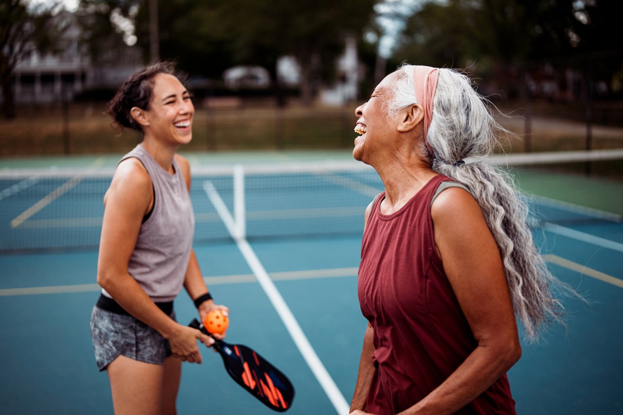 A senior woman and her adult daughter laugh together on an outdoor pickleball court after a match.
