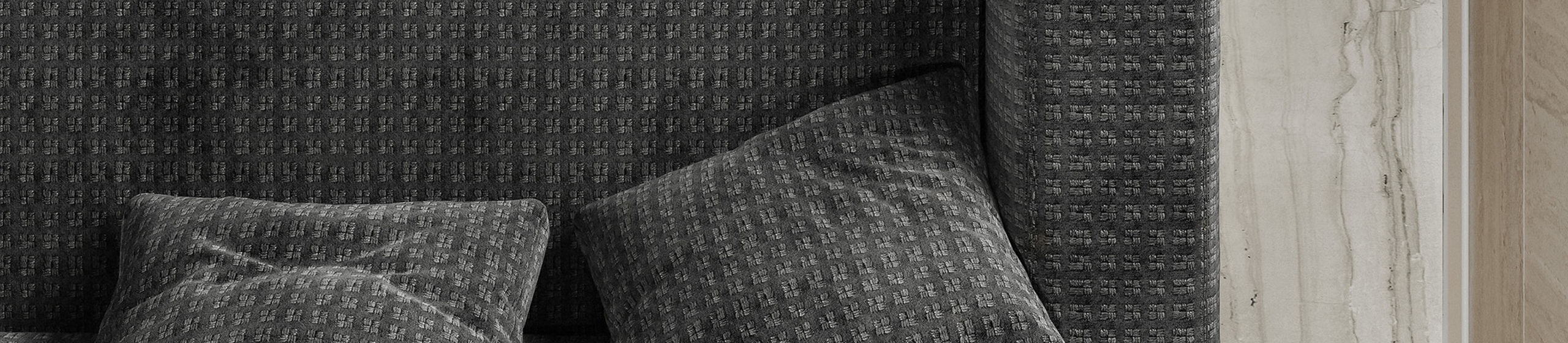 A contemporary gray upholstered sofa is positioned in front of a light marble-tiled wall. Two matching gray cushions rest casually on the seat, adding texture and comfort. The marble wall features subtle vertical veining in beige and taupe tones. A light wood cabinet or panel is partially visible on the right, contributing to the minimalist interior design.