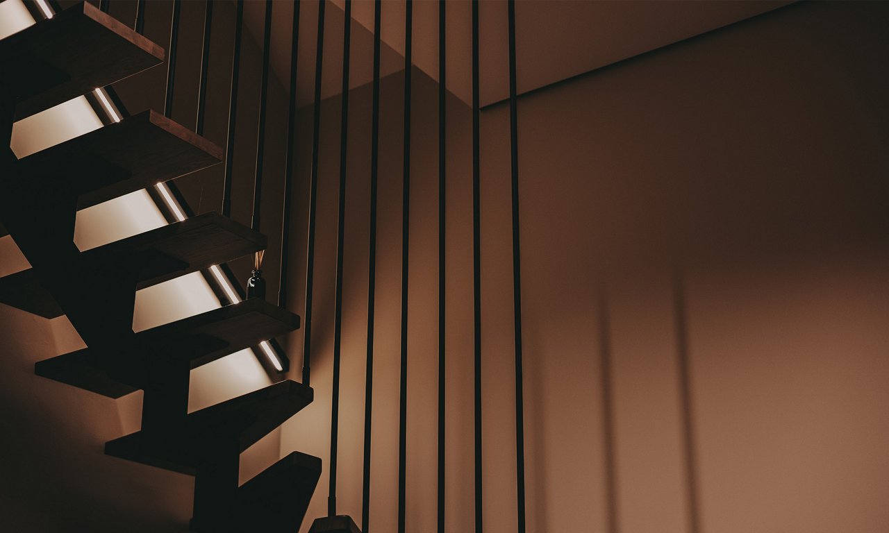 A modern floating staircase with dark wooden treads is shown from a low, angled perspective. Slim vertical metal railings extend from the steps to the ceiling, creating a strong linear pattern. Warm ambient lighting casts soft shadows on the smooth walls, emphasizing the minimalist architecture. The overall scene feels contemporary and refined, highlighting clean lines and geometric forms.