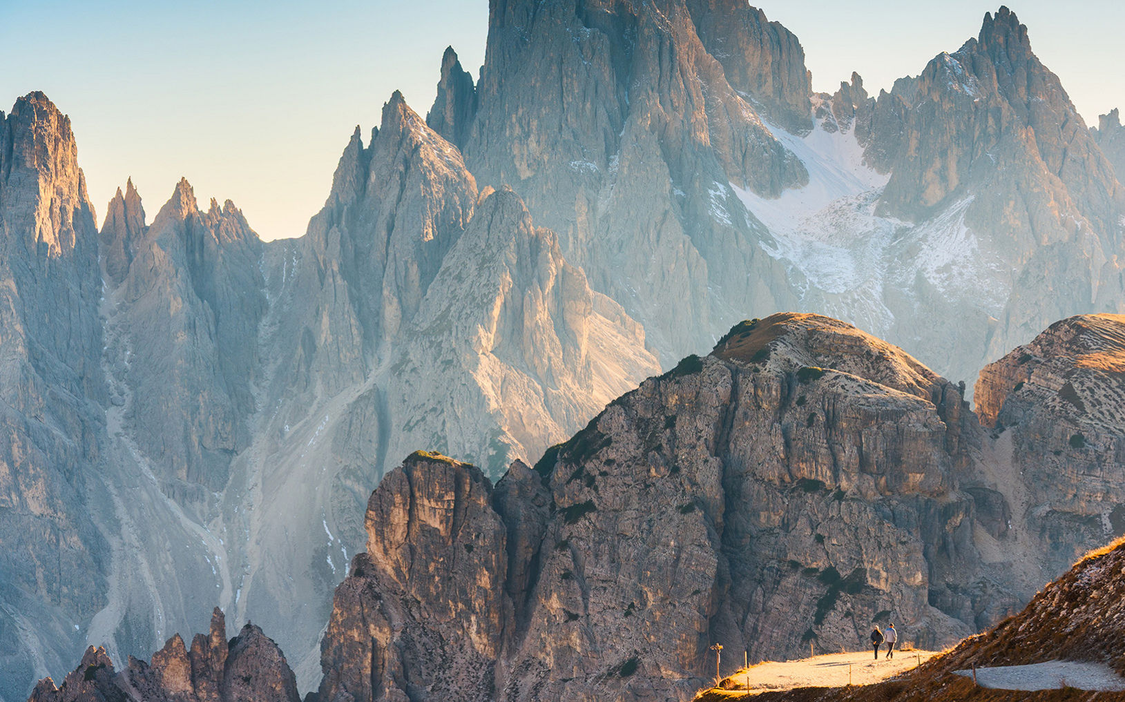 Two hikers walk along a narrow sunlit trail high in a rugged mountain range. Towering jagged peaks rise steeply in the background, some dusted with snow in shaded gullies. Warm golden light from the low sun illuminates the foreground slopes, contrasting with the cool blue tones of distant rock faces. The expansive landscape emphasizes scale and adventure in a remote alpine setting.