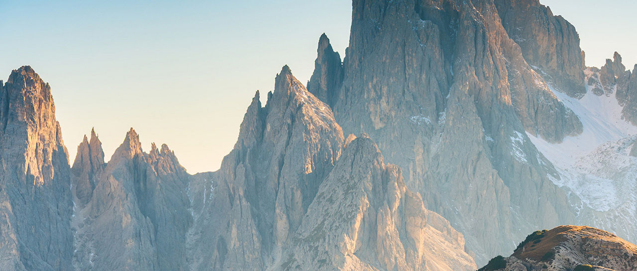 Two hikers walk along a narrow sunlit trail high in a rugged mountain range. Towering jagged peaks rise steeply in the background, some dusted with snow in shaded gullies. Warm golden light from the low sun illuminates the foreground slopes, contrasting with the cool blue tones of distant rock faces. The expansive landscape emphasizes scale and adventure in a remote alpine setting.