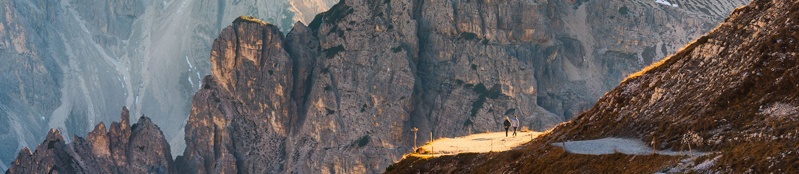 Two hikers walk along a narrow sunlit trail high in a rugged mountain range. Towering jagged peaks rise steeply in the background, some dusted with snow in shaded gullies. Warm golden light from the low sun illuminates the foreground slopes, contrasting with the cool blue tones of distant rock faces. The expansive landscape emphasizes scale and adventure in a remote alpine setting.