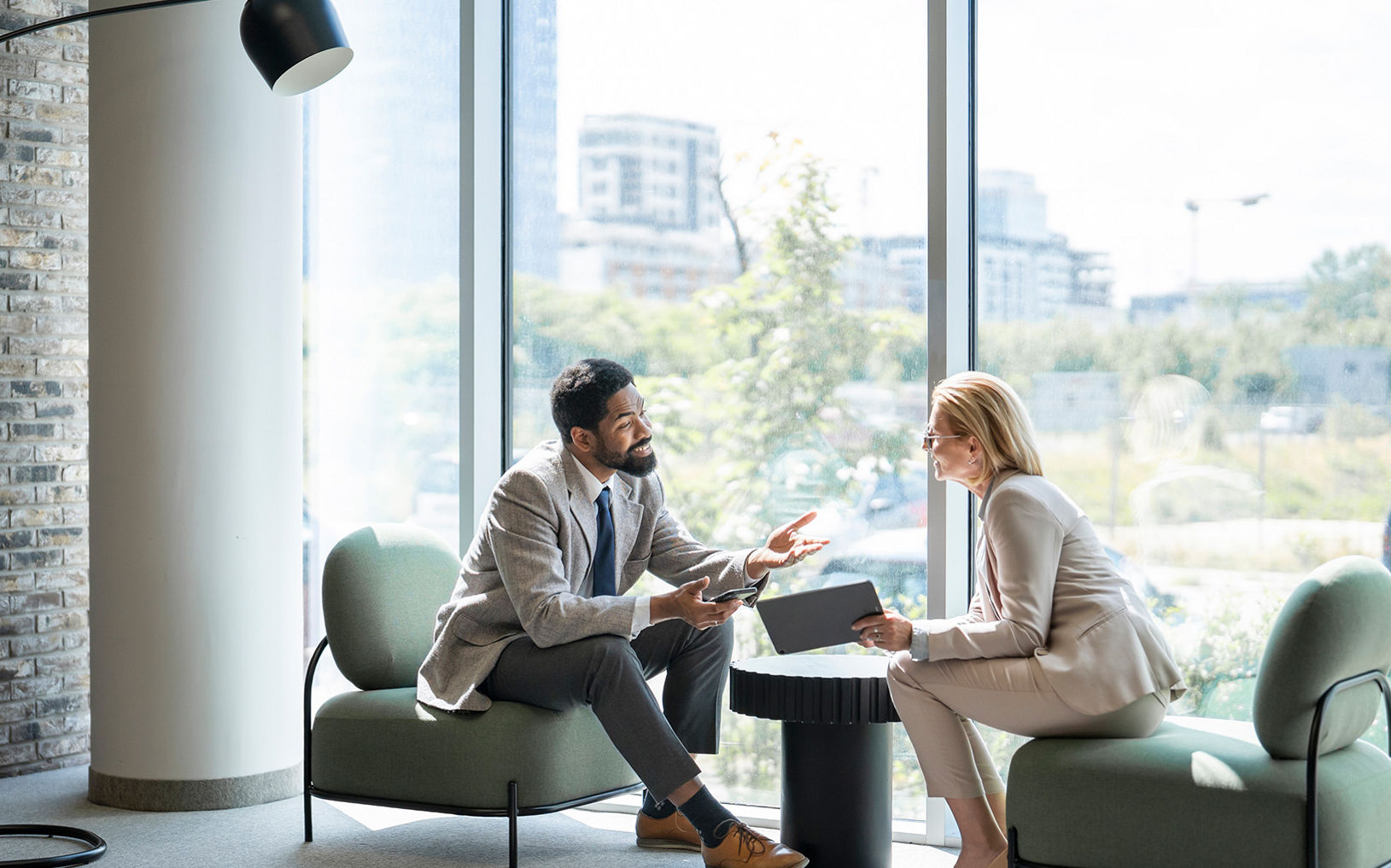 Two business professionals sit facing each other in a modern office lounge area. They are engaged in a discussion, with one person holding a digital tablet and the other gesturing while speaking. Large floor-to-ceiling windows reveal an urban cityscape and allow natural light to fill the space. Contemporary furniture and a tall floor lamp contribute to the clean, professional atmosphere.
