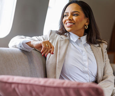 Une femme en tenue professionnelle est confortablement installée dans un siège moelleux à l’intérieur de la cabine d’un avion privé. Elle porte un blazer clair par-dessus une chemise blanche et son bras est posé nonchalamment sur l’accoudoir. Les coussins moelleux et les tissus d’ameublement aux tons neutres contribuent à créer une atmosphère raffinée et haut de gamme. La lumière naturelle pénètre par le hublot de l’avion, mettant en valeur un moment de calme et de détente lors d’un voyage d’affaires.