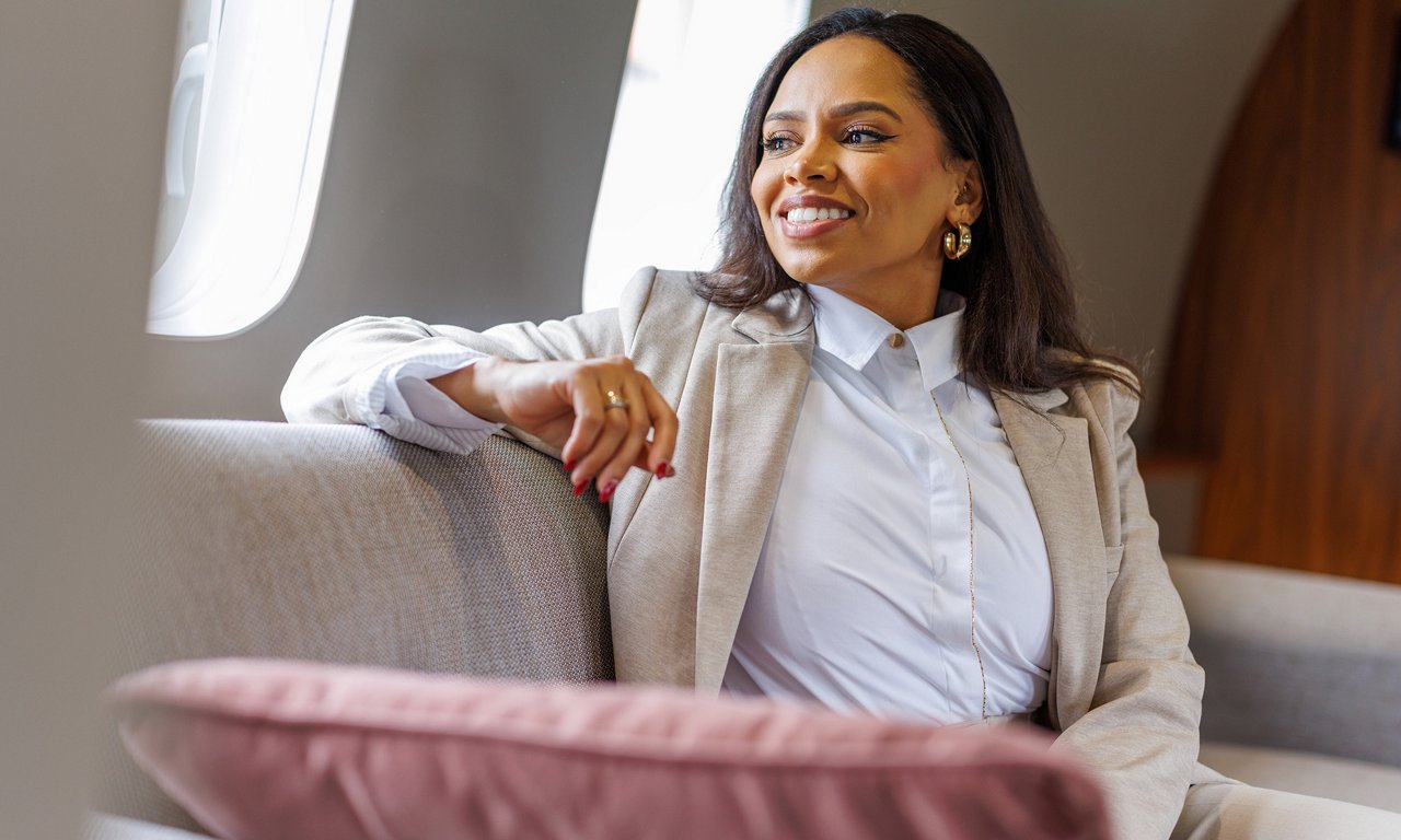 A professionally dressed woman sits comfortably on a plush seat inside a private airplane cabin. She wears a light-colored blazer over a white shirt and rests her arm casually on the armrest. Soft cushions and neutral-toned upholstery contribute to a refined, upscale atmosphere. Natural light enters through the aircraft window, highlighting a calm and relaxed business travel moment.