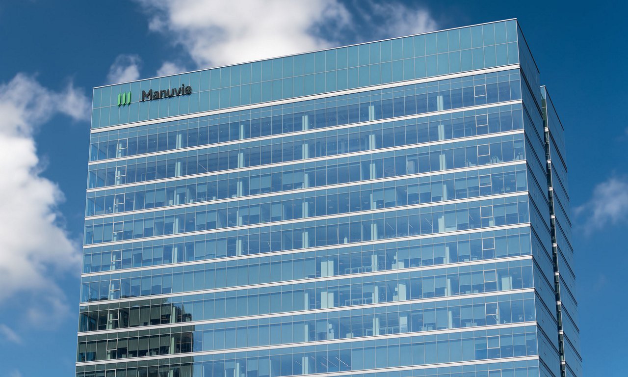 A tall modern office building with a glass facade rises against a blue sky with scattered clouds. The structure features multiple floors with large reflective windows and visible interior workspaces. A green and white corporate sign with the word "Manulife" appears near the top of the building. The overall scene conveys a contemporary urban business environment and corporate headquarters setting.