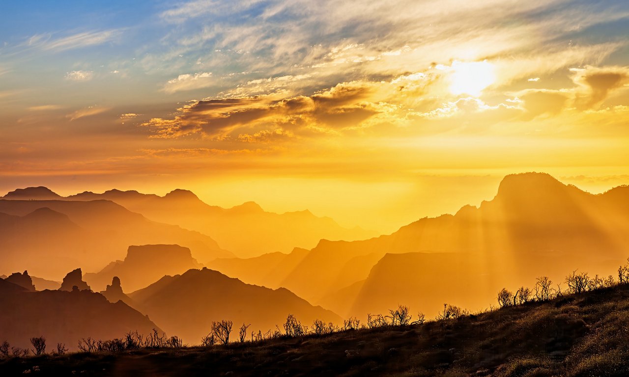 A dramatic sunrise casts golden light over a layered mountain range. Soft mist fills the valleys, creating a dreamy, atmospheric depth between the ridges. Sunbeams break through scattered clouds, illuminating the landscape with warm, glowing tones. Dry grasses in the foreground add texture and contrast against the bright sky.