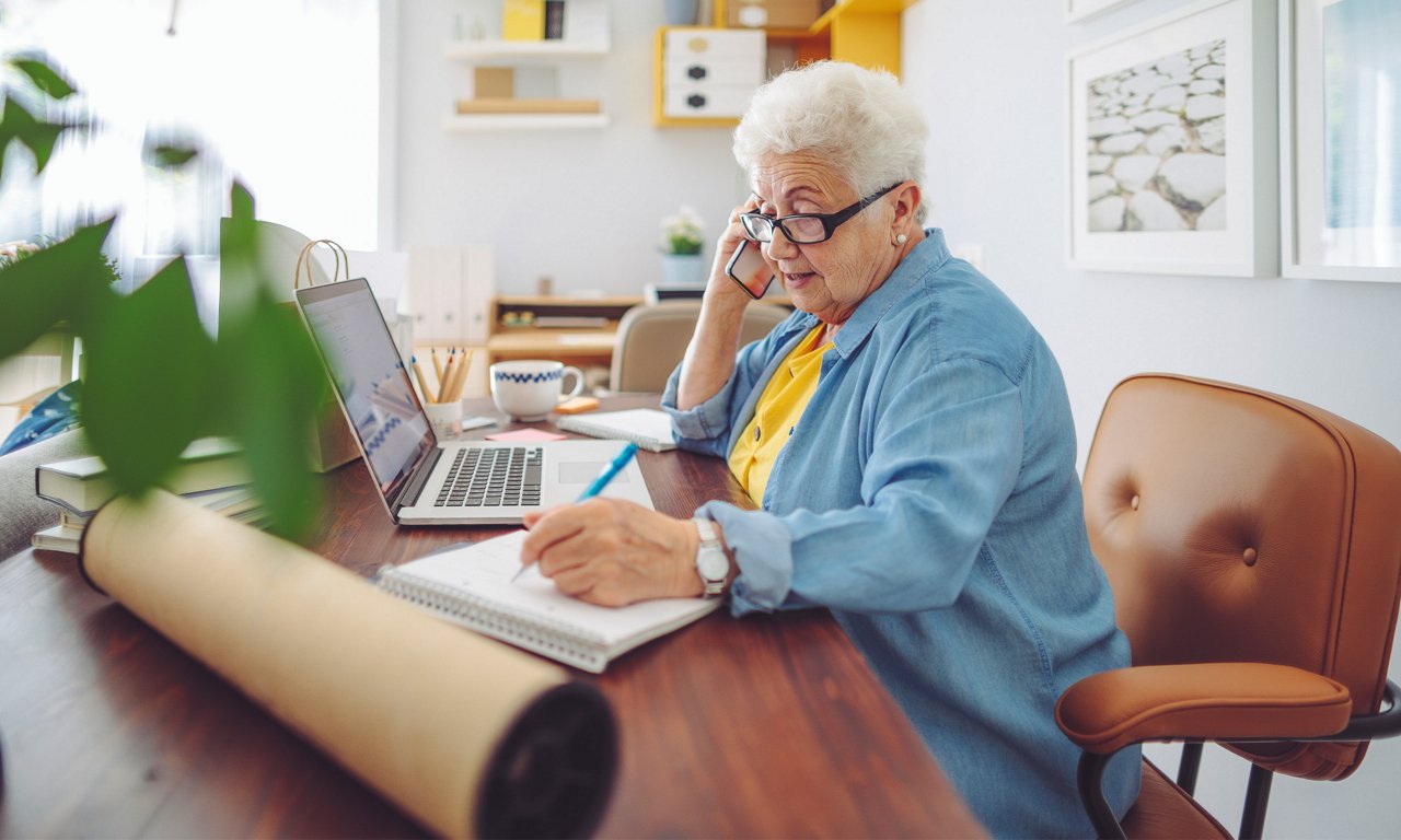 Older woman reviewing information on a laptop while talking on the phone and taking notes at home.