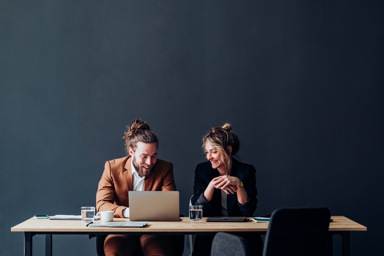Happy businessman and businesswoman using a laptop computer while talking and sitting together at office desk.