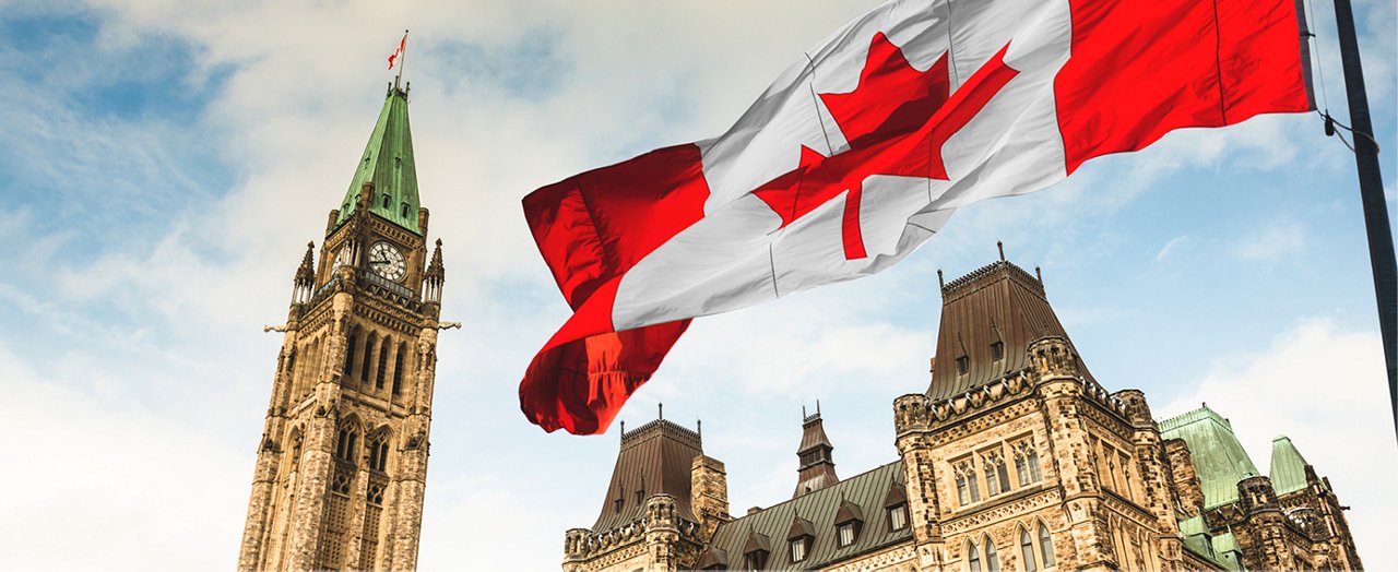 The Canadian government's flag majestically flies against the blue sky over a European city's architectural tower.