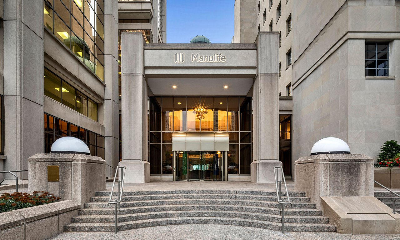 A modern corporate office entrance features the Manulife name above a glass doorway. Wide stone steps and metal railings lead up to the main doors, framed by tall concrete and glass facades. Symmetrical round lights and planters with flowers accent the foreground. Warm interior lighting contrasts with the cool exterior tones, creating a professional and welcoming atmosphere.