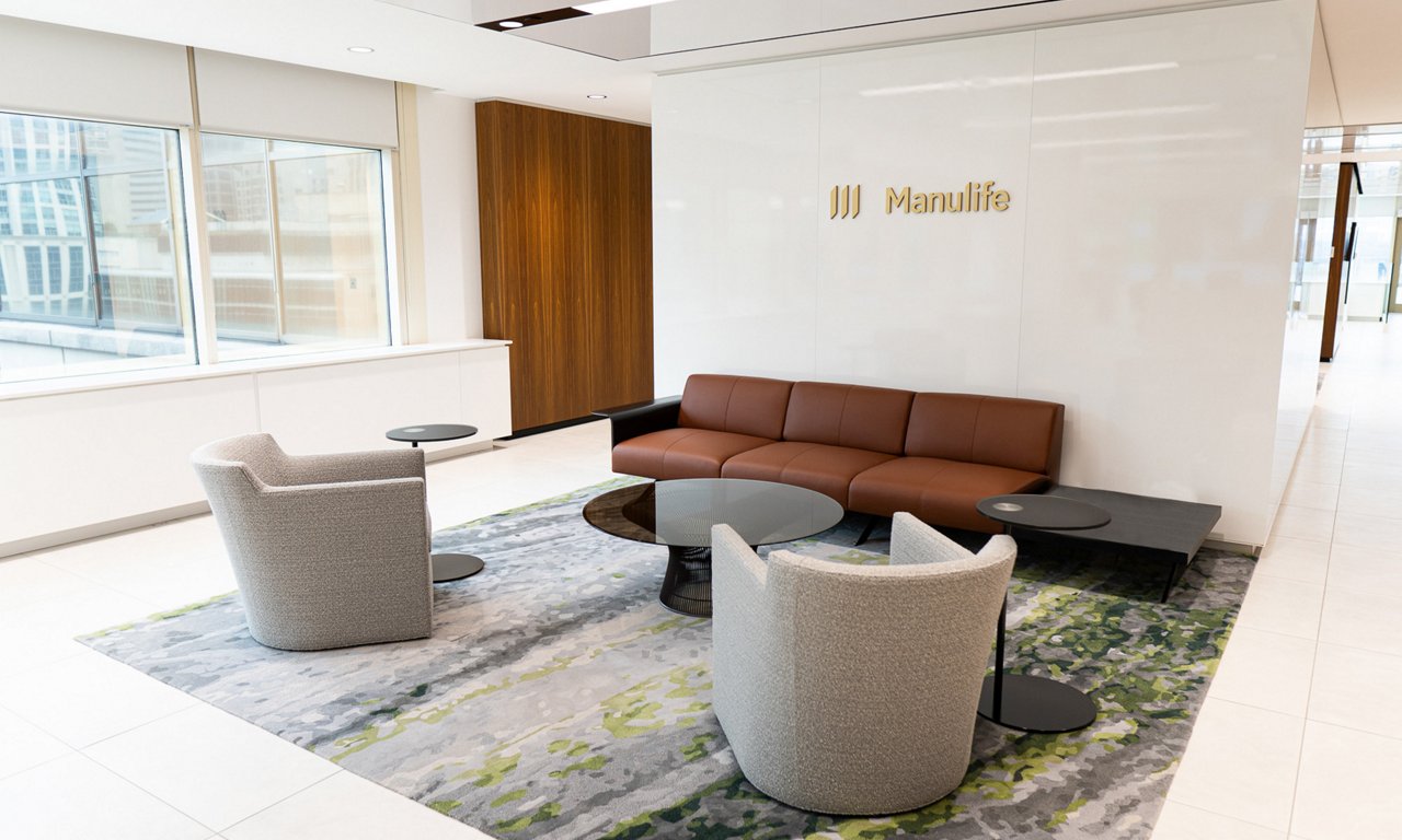 A contemporary office lobby features a brown leather sofa and two light gray lounge chairs arranged around low black coffee tables. The seating area sits on a patterned area rug with green and gray tones, contrasting with the white tile floor. Large windows on the left bring in natural light and reveal an urban cityscape outside. The Manulife name and emblem are prominently displayed on a clean white wall behind the sofa.
