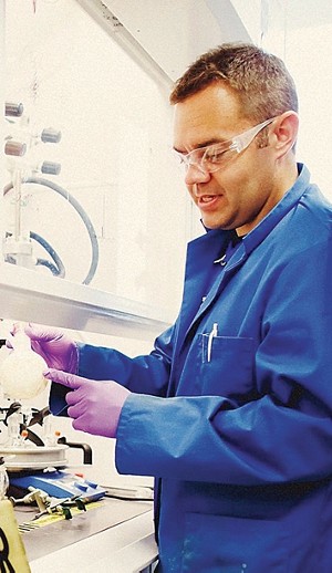 A scientist, Simon Humphrey, wearing protective eyewear and a lab coat in the lab, pointing to a white balloon that is filling with something, probably a gas.