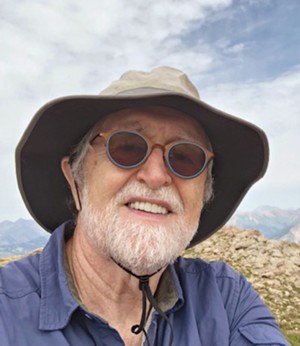 Photo of Frederick Turner wearing a floppy hat and sunglasses hiking under a cloudy sky.