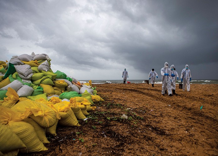 Under heavy skies, a group of people in white full-body protective gear walk along a beach. Plastic bags are piled along the beach