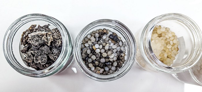 Photo looks into the mouths of three jars. The jar on the left holds agglomerated, charred pieces of plastic; the jar in the middle holds sooty grey plastic pellets; the jar on the right holds orange and off-white plastic pellets.