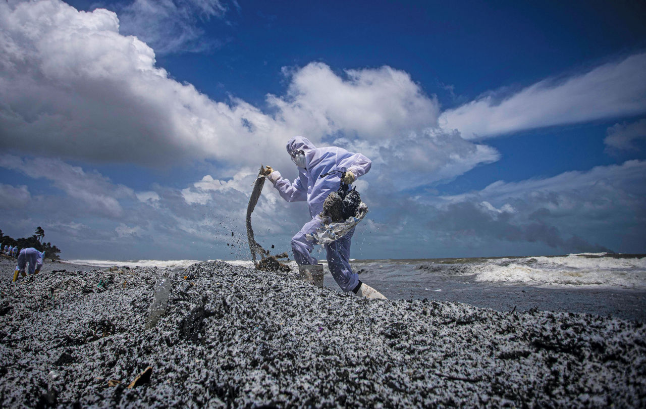 A person clad in full-body protective suit walks on mounds of white and black plastic debris on a beach. Behind the person, a wave washes to the shore and clouds mass in a blue sky.