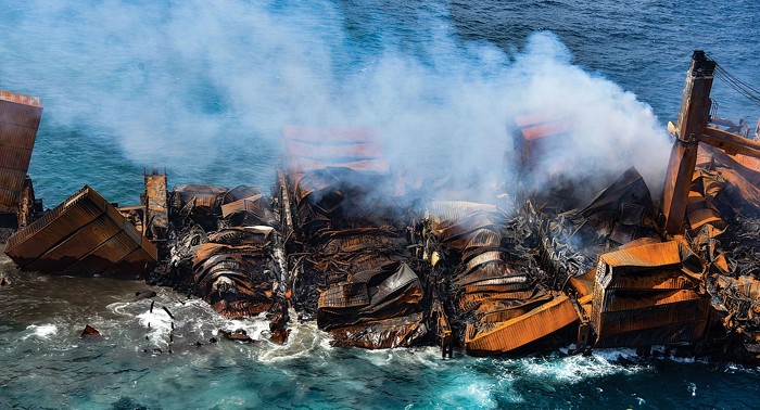 Photo shows the top part of a sinking ship, just above water. Smoke pours from the ship and twisted cargo containers are visible.