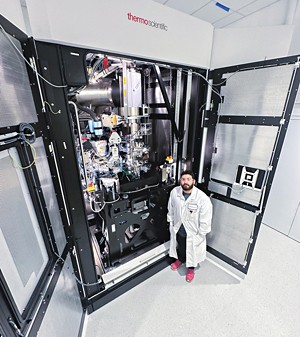 A man in front of a microscope twice as tall as he is and about as deep as a walk-in closet, façade open to show a tangle of tubes and parts.