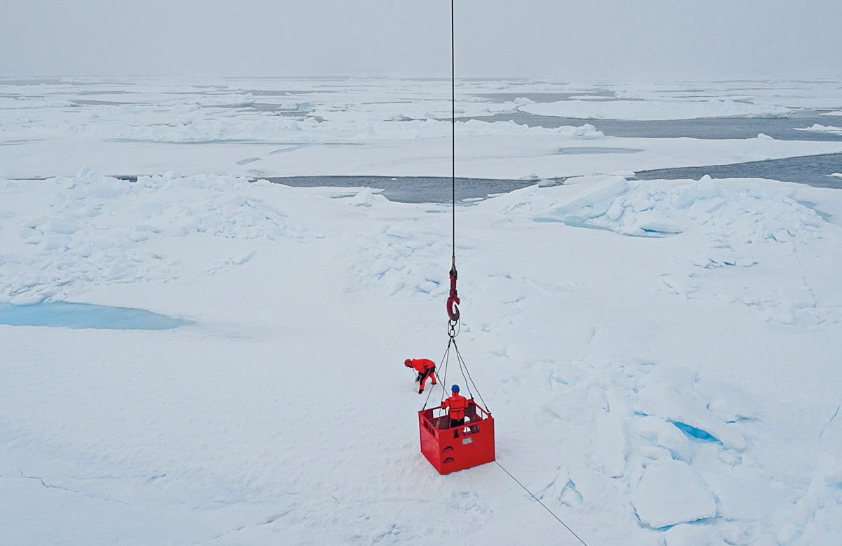 Two people in orange gear. One of them is being lowered onto the snow by a crane.