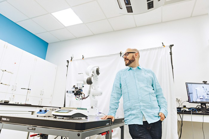 A man standing in a laboratory setting between a robotic device and a computer screen.