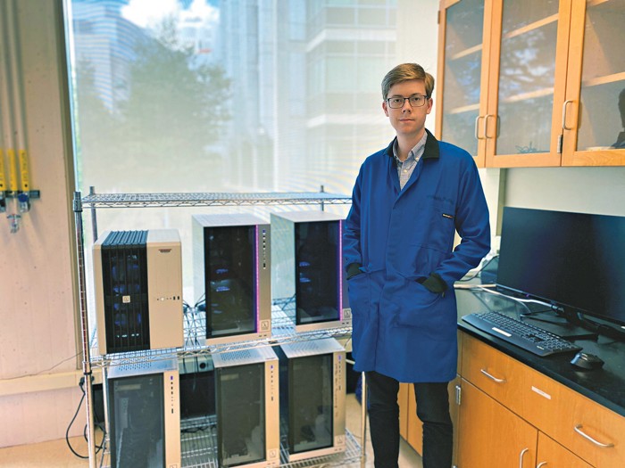 A man in a lab coat and goggles next to computer equipment.