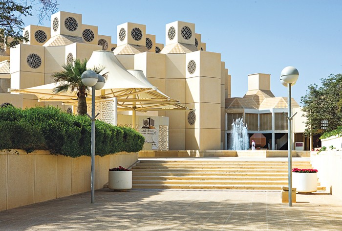 In the foreground is a courtyard paved with pale-yellow tiles. Steps lead to a fountain and a similarly colored building with a blocky, angular structure. Several cubes sit atop the building, and each cube’s horizontal side includes an ornate screen.