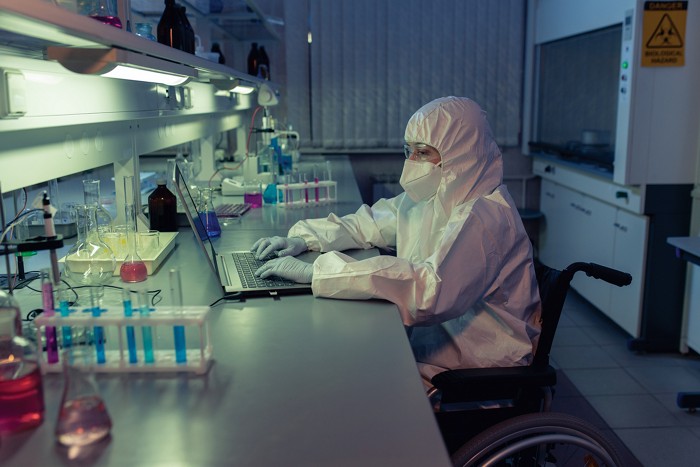A person sitting in a wheelchair at a laboratory bench is typing on a laptop. In the foreground are beakers and vials filled with liquids of different colors. The researcher is wearing a face mask, safety goggles, gloves, and a white protective suit.