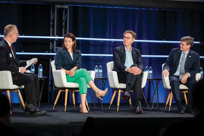 Sitting on a brightly lit stage are four people. The person on the left is holding a piece of paper, and the three others are turned toward him, listening.