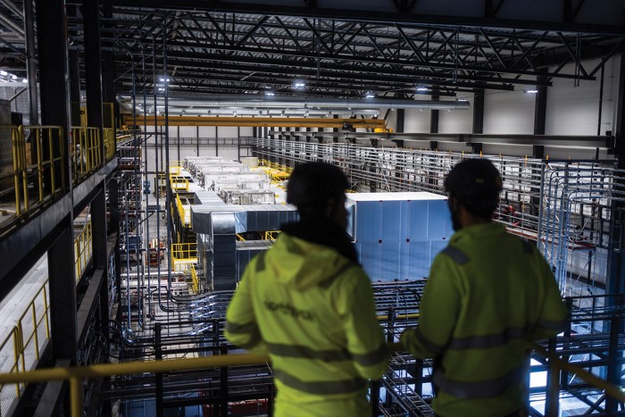 Two workers in fluorescent yellow jackets stand on the upper level of a large warehouse filled with equipment for recycling batteries.